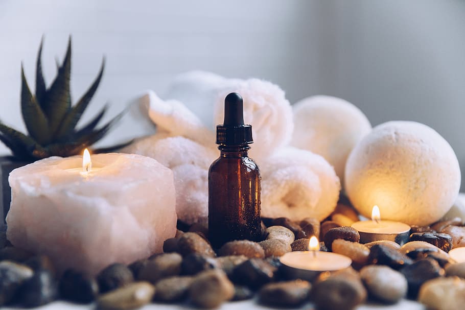 Photo of a brown vial of essential oil settled into smooth rocks and surrounded by a softly glowing candle, succulent, bath bombs, and towels.
