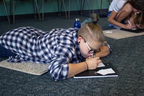 Photo of a teen wearing a flannel shirt, laying on the floor and writing in a notebook.