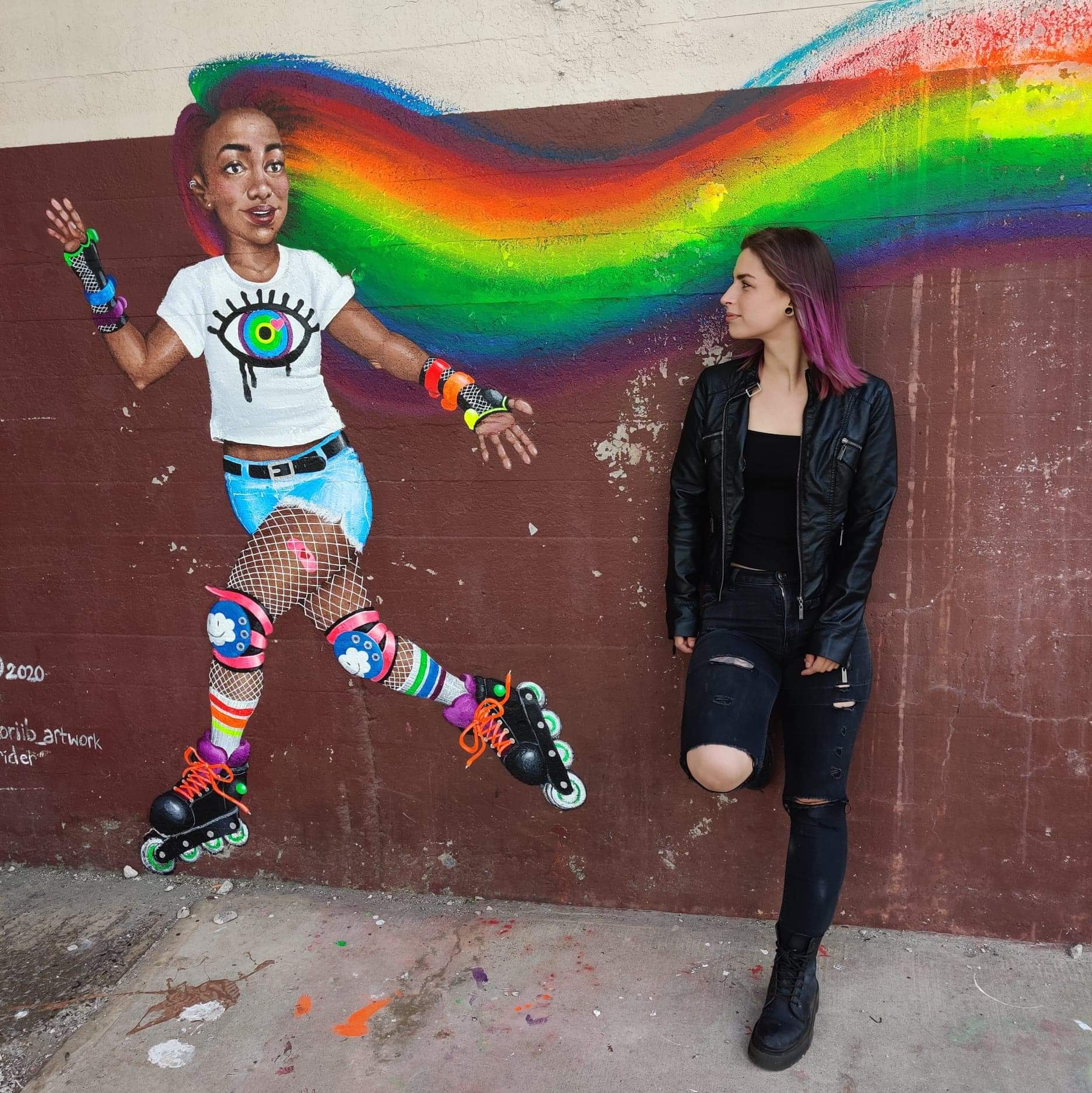 Photo of a young woman with purple hair and light skin looking at a painting of a teen girl with dark skin who's hair transforms into a rainbow.