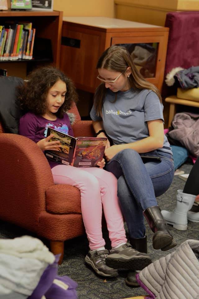 A young woman reading a book with a younger child, seated in a chair.