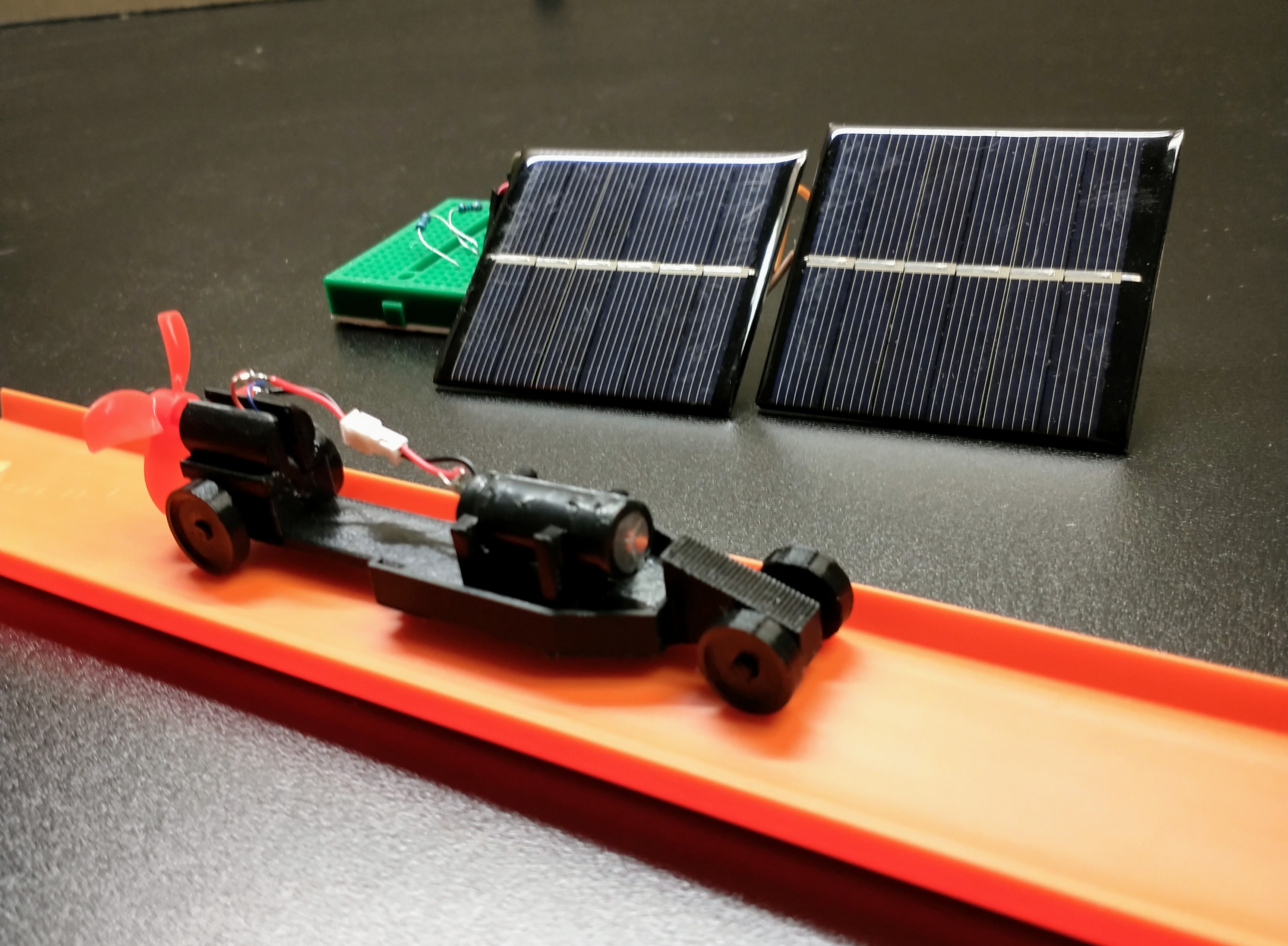 A 3D printed car on orange Hot Wheel track next to two solar cells.