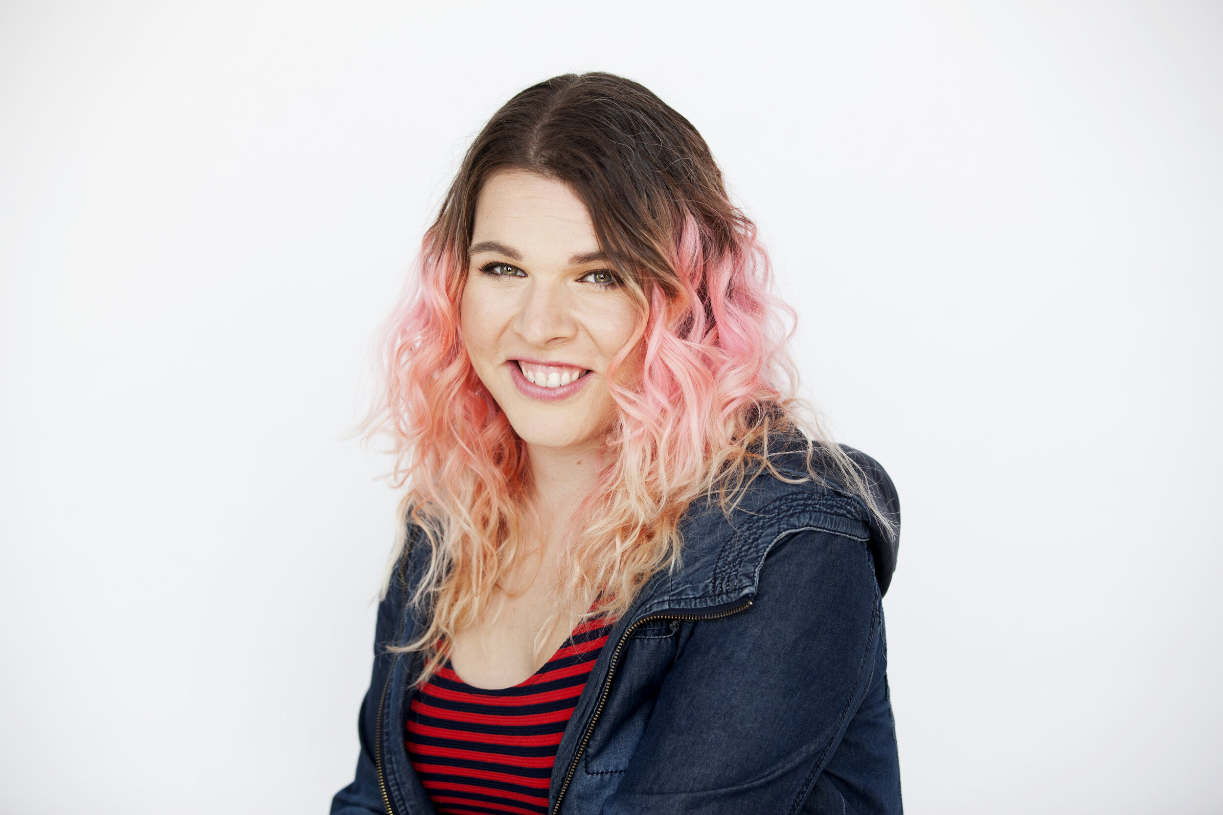 Photo of a fair-skinned woman with long wavy hair that is dark at the roots and pink halfway down. She is smiling and wearing a denim zip up hoodie and a black and red striped shirt.