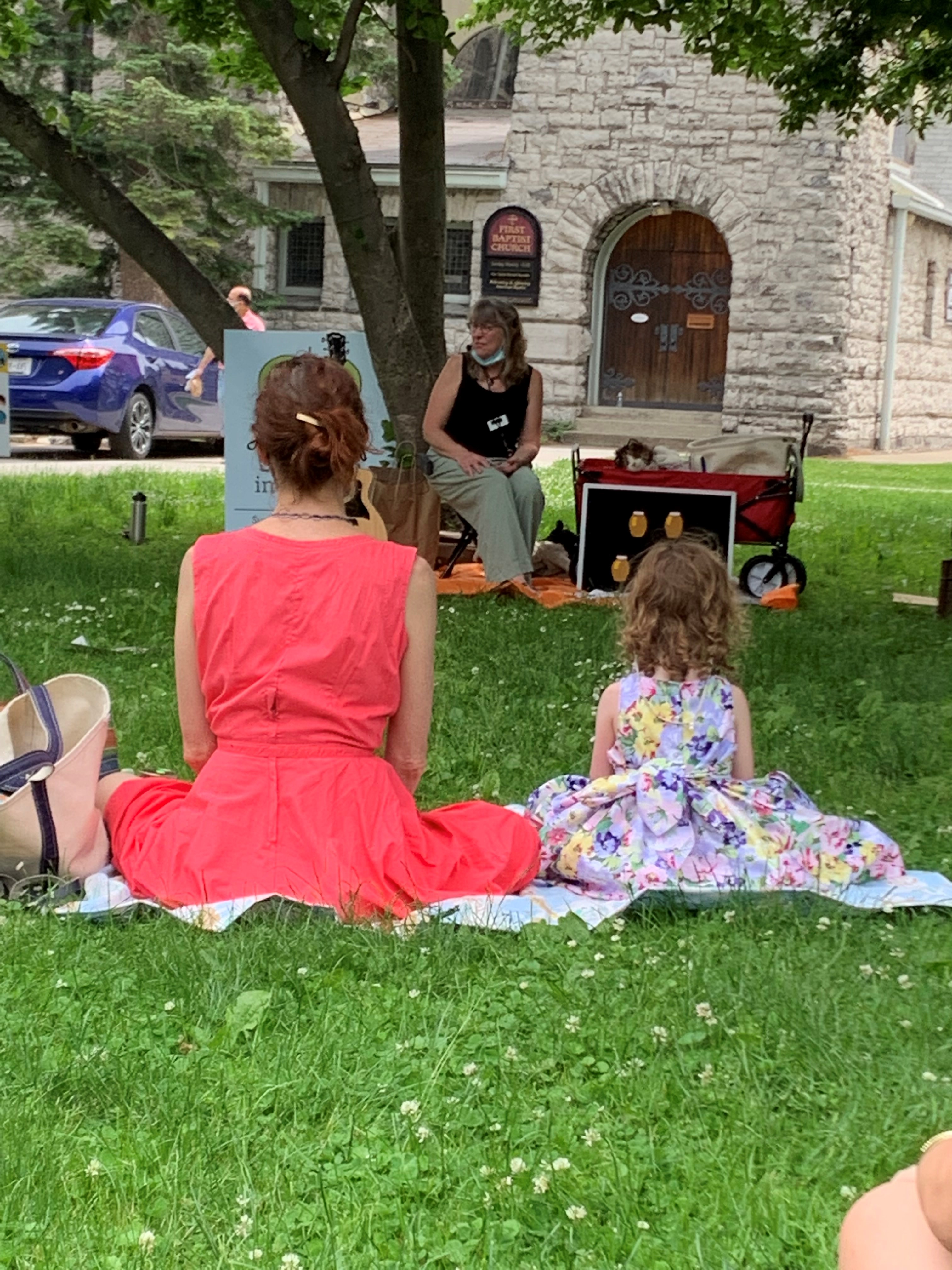 Mother in pink dress sitting next to young girl in floral dress in the park, listening to storytime.