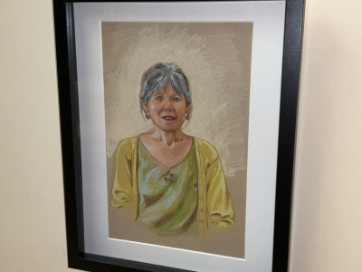 A colored-pencil portrait by Yamilka Pororreal showing Betty Spero. In the portrait Betty is smiling, and wearing a green shirt with a floral design at the center, and is also wearing a yellow cardigan sweater.