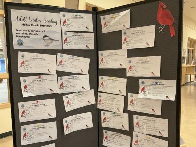 A gray standing poster board, with a dozen or more small pieces of paper attached. Each contains a haiku about a book. There is an image of a bright red cardinal on the top right hand corner of the poster board, and a sign reading "Adult Winter Reading - Haiku Book Review" on the top left corner