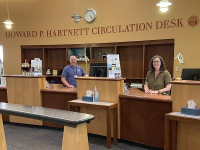 Librarians Tom (left) and Kate (right) at the Circulation Desk. It is a large wooden desk, with a tan-colored wall behind it that has a sign identifying it as the Howard P. Hartnett Circulation Desk.
