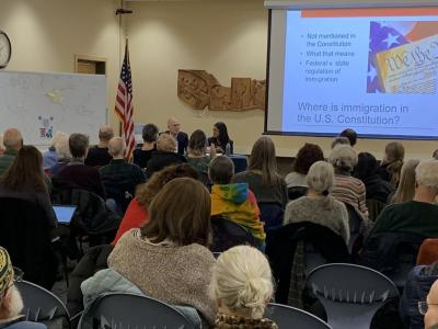 An audience in the BorgWarner Community Room watching Steve Yale-Loehr and Tania Penafort speak. To the right is a presentation on a screen. Behind them, an American flag is visible. On the left is a world map that audience members marked with their family's country of origin.