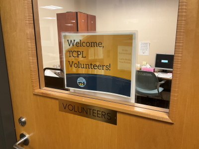 The door to the Volunteer room at TCPL. It is a wooden door that has a large window on it. A brass plate reads "Volunteers" and there is a sign in the window that says WELCOME TCPL VOLUNTEERS.