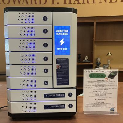 The phone charging locker. It is a big white, rectangular shaped metal box that has six small locker-style doors on the left, a touchscreen on the right, and two large doors at the bottom.