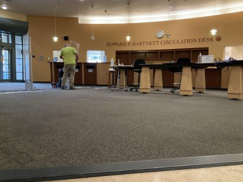 A wide image of the TCPL lobby, showing men putting a partition into place for the RFID gates. The Howard P. Hartnett Circulation Desk and sign are visible in the background.
