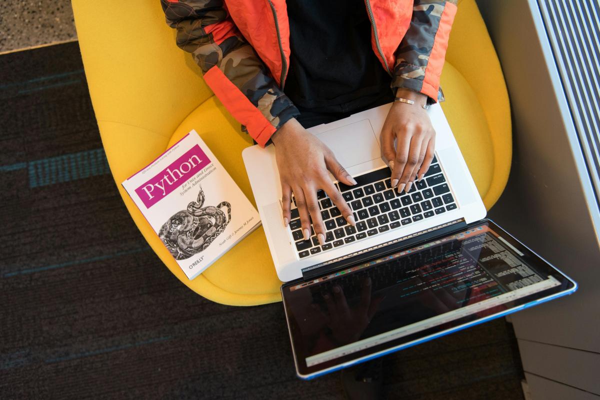 Person writing python scripts on computer with text book next to their laptop.