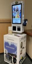 The Ezra Coin-ell donation kiosk. It is a large white metal box, with a vertical touch screen monitor attached to the top. It includes pictures of various TCPL events, and has a credit card reader on the right hand side.