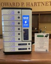 The phone charging locker. It is white metal, with a touch screen panel on the right hand side that reads "charge your device here. Touch to begin." On the left side are six locker doors. Two large locker doors are located at the bottom.