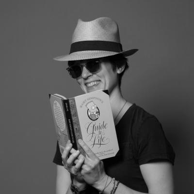 A picture of youth librarian Kat wearing a fedora style hat and sunglasses. She is reading a book titled "Bart Simpson's Gide to Life"